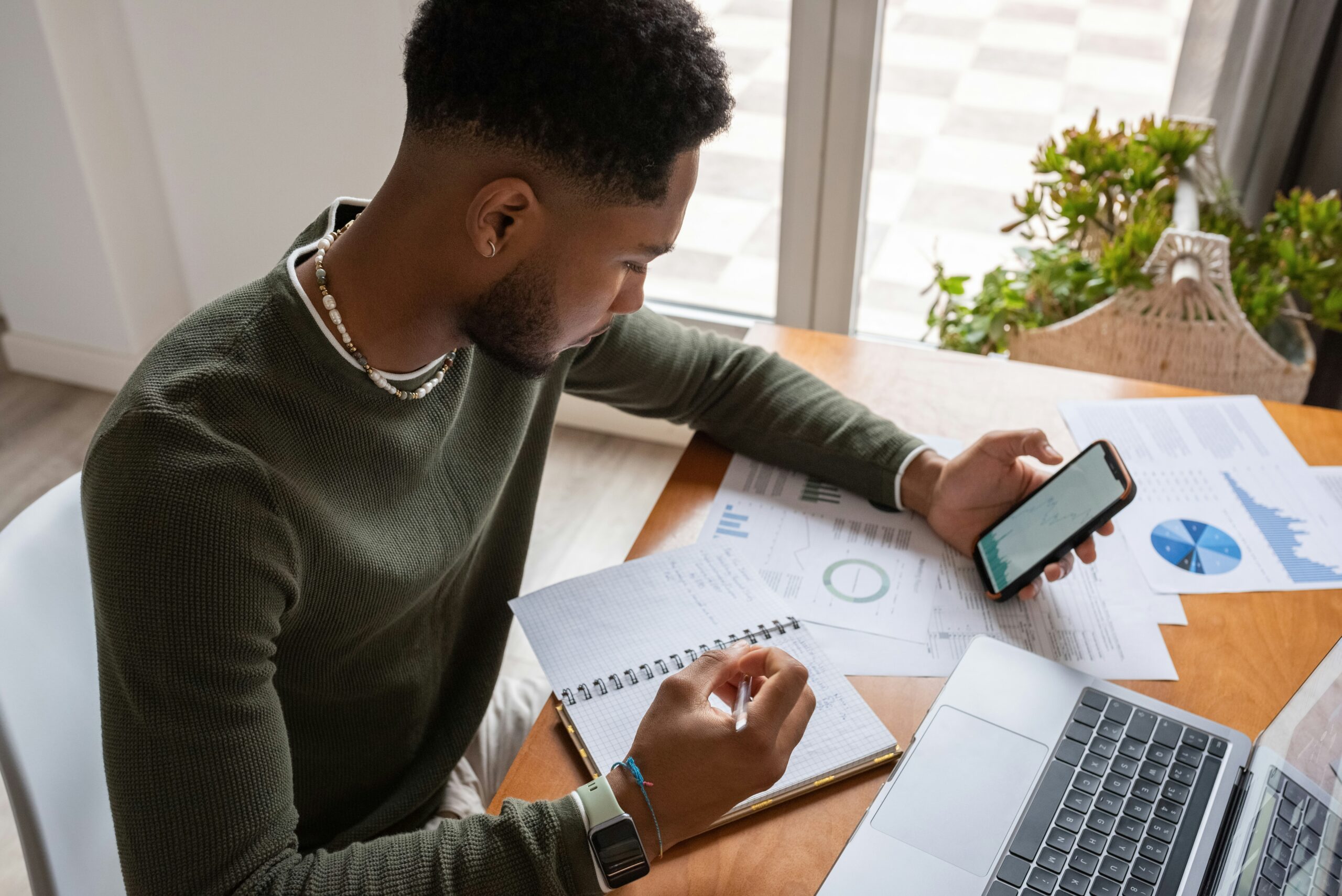 Young man working on his finances at a desk with his phone and laptop, making notes in a notebook